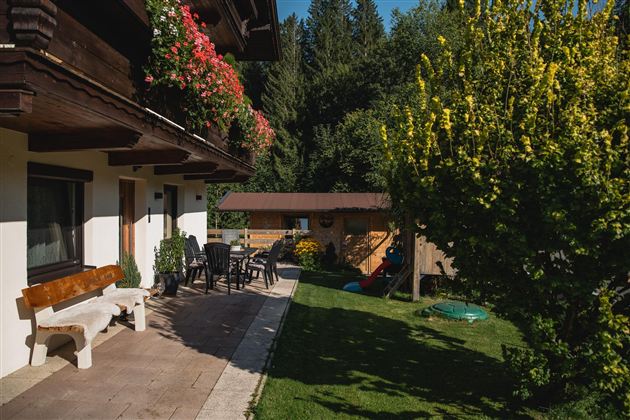Ein gemütlicher Außenbereich mit einer Terrasse, Tisch und Stühlen. Im Hintergrund sind Bäume und ein Holzhaus sichtbar.