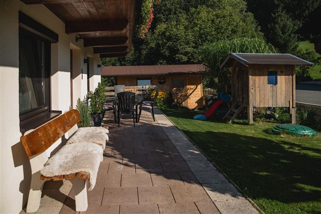 Eine einladende Terrasse mit einer Bank und Stühlen. Im Hintergrund befindet sich ein Gartenhäuschen und Spielgeräte auf der Wiese.