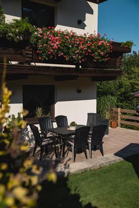 Ein schöner Außenbereich mit einem Tisch und Stühlen aus Kunststoff. Blumen hängen am Balkon und der Rasen ist grün und gepflegt.