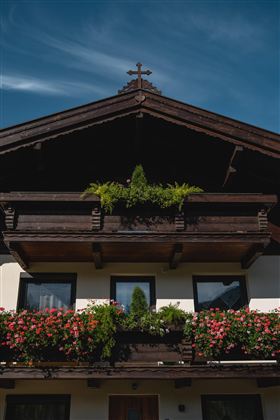 Ein traditionelles alpenländisches Haus mit Holzbalkonen und bunten Blumen. Darüber erhebt sich ein Kreuz auf dem Dach unter einem klaren blauen Himmel.