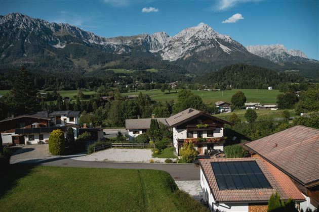 Eine malerische Landschaft mit grünen Wiesen und charmanten Häusern. Im Hintergrund erheben sich beeindruckende Berge unter einem klaren blauen Himmel.