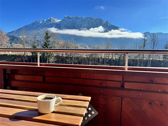 Eine gemütliche Terrasse mit einem Holz Tisch und einer Tasse Kaffee. Im Hintergrund sind majestätische Berge und ein klarer blauer Himmel zu sehen.