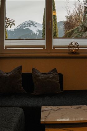 A cozy living room with a dark gray sofa and cushions. A snow-covered mountain can be seen through the window.