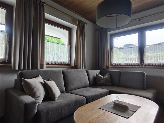 A cozy living room with a gray sofa and a wooden table. The windows are adorned with curtains and provide a view of the outdoors.