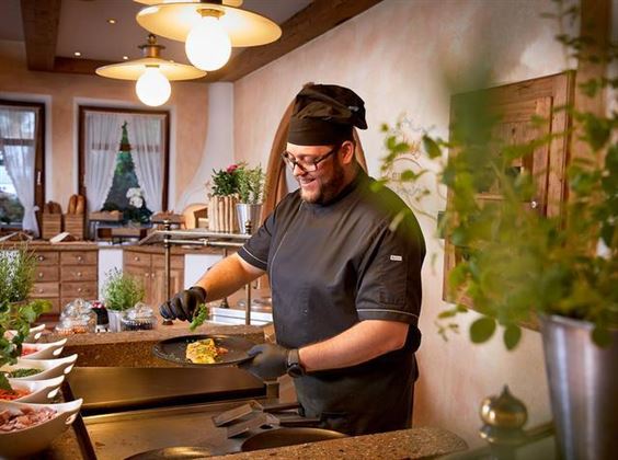 A cook is preparing a meal in a cozy kitchen. The environment is bright and friendly, with plants and decorative elements.