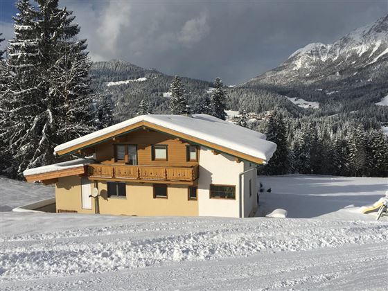 A cozy house in a snow-covered landscape, surrounded by pine trees. In the background, gentle hills and mountains can be seen.