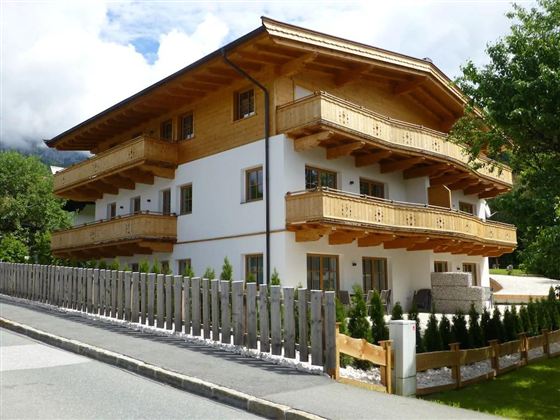 Ein modernes Haus im alpenländischen Stil mit mehreren Balkonen. Es ist von einem gepflegten Garten und einem Zaun umgeben.