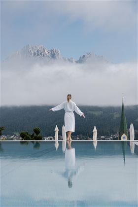 Een vrouw in een badjas staat aan de rand van een infinity-pool. Op de achtergrond zijn bergen en mist te zien, wat een rustige en ontspannen atmosfeer creëert.