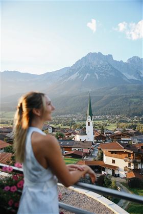 Een vrouw staat op een balkon en kijkt uit op een pittoreske stad met een kerk en bergen op de achtergrond. De zon schijnt en creëert een warme sfeer.