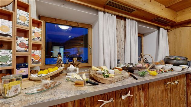 A buffet with various types of cheese, cold cuts, and fresh fruit. The room has large windows with a view outside and a cozy wooden structure.