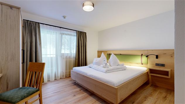 A bright hotel room with a double bed and a wooden desk. In front of the window are long curtains and the floor is made of wood.
