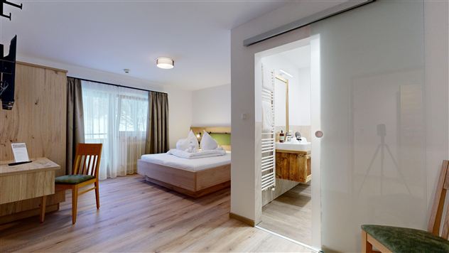 A modern hotel room with a large bedroom and a bathroom. The space is bright and friendly decorated, with wooden furniture and a window.