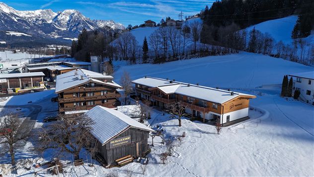 A picturesque snowy landscape with several wooden houses in the foreground. In the background, the snow-covered mountains and a blue sky can be seen.
