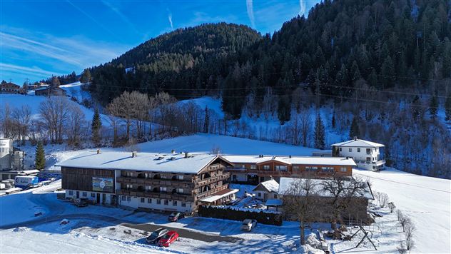 A rustic hotel in a snow-covered landscape. In the background, forested mountains and a blue sky can be seen.