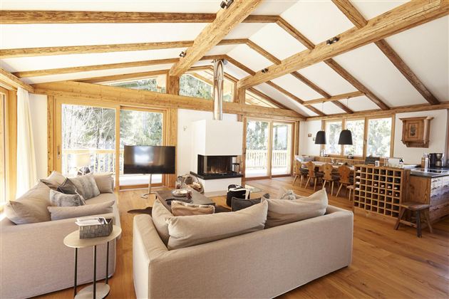 Ein modernes Wohnzimmer mit Holzdecken und großen Fenstern. Es gibt gemütliche Sofas, einen Esstisch und einen Kamin.