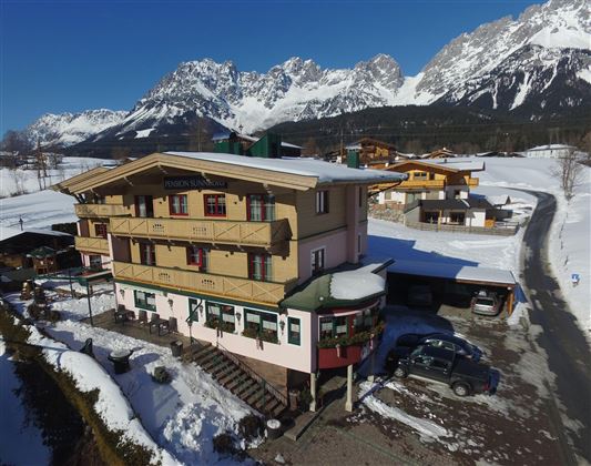 A cozy hotel in the mountains, surrounded by snow-covered landscapes. The majestic mountains rise in the background under a clear blue sky.