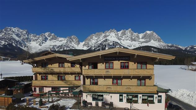 A cozy chalet in the mountains with snow-covered peaks in the background. Clear blue skies and a tranquil setting.