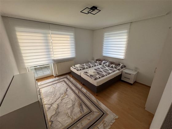 Ein helles Schlafzimmer mit zwei Betten und einem modernen Teppich. Große Fenster lassen viel Licht herein und sorgen für eine einladende Atmosphäre.