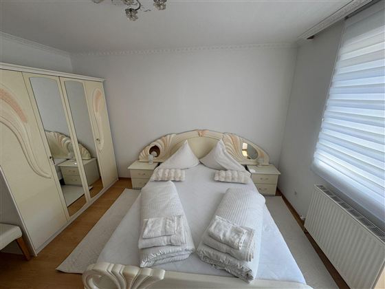 A bright bedroom with a large bed and beige walls. There is a mirror and a large wardrobe in the room.