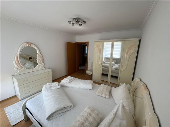 A bright bedroom with a large bed and an elegant dresser. The room is simply and modernly furnished, with a large mirror and a spacious wardrobe.