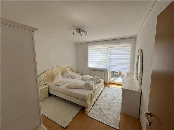 A bright bedroom with a large bed and white bedding. There is a large window with blinds and a mirror on the wall.