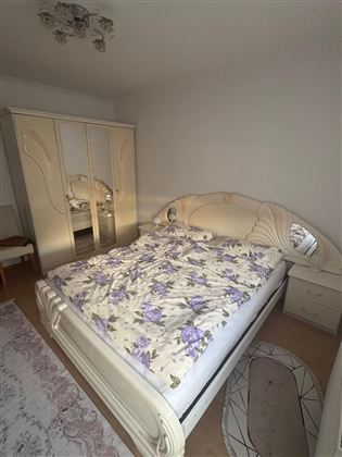 Ein helles Schlafzimmer mit einem großen Bett und einer weißen Bettwäsche, die mit lila Blumenmuster verziert ist. Im Hintergrund steht ein großer Kleiderschrank mit Spiegel.