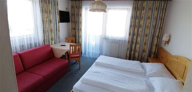 Een gezellige kamer met een tweepersoonsbed en een rode bank. Grote ramen zorgen voor veel natuurlijk licht.