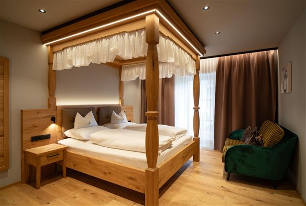 A cozy bedroom with a wooden four-poster bed. There is a small table and a green couch, as well as large windows with curtains.