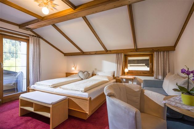 Ein gemütliches Schlafzimmer mit einem Doppelbett und einer Sitzecke. Der Raum hat helle Wände und ein großes Fenster, das Tageslicht hereinlässt.