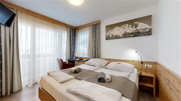 A bright hotel room with a double bed and wooden furniture. Large windows offer a beautiful view of the mountains.