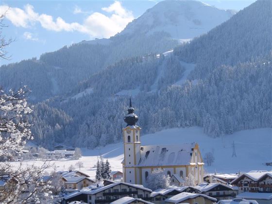 Eine malerische Winterlandschaft mit schneebedeckten Bergen. Im Vordergrund steht eine schöne Kirche umgeben von traditioneller Architektur.