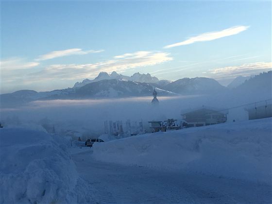 A snow-covered landscape with mountains in the background and a misty sky. The shoreline is covered in snow and it is very quiet.