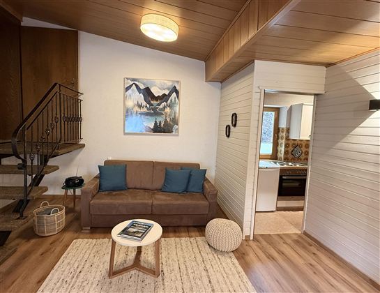 Ein gemütliches Wohnzimmer mit einem Sofa, zwei Kissen und einem kleinen Tisch. An der Wand hängt ein Bild mit einer Berglandschaft.