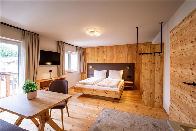 A modern room with wooden decor, a double bed, and a table. Large windows provide plenty of daylight and a cozy atmosphere.