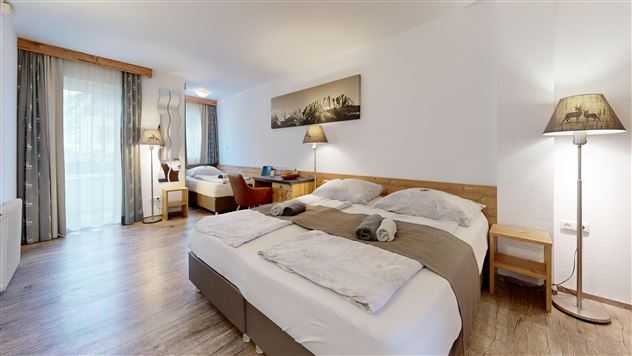 A cozy hotel room with a large double bed and wooden furniture. The windows provide daylight and there are decorative lamps as well as a picture on the wall.