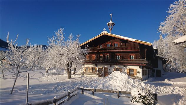 Een traditioneel houten huis in een besneeuwde landschap. Omringd door met sneeuw bedekte bomen en een heldere, blauwe lucht.