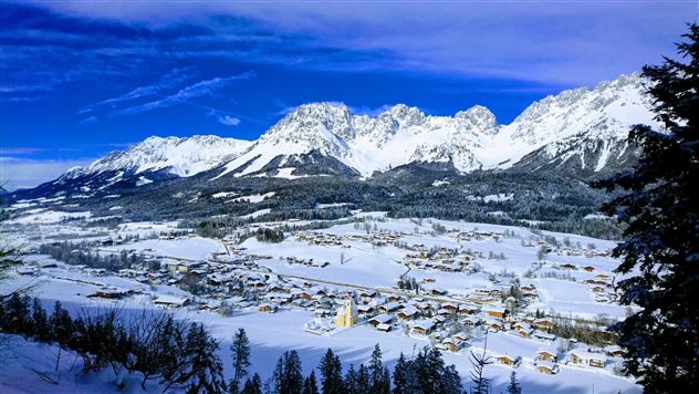 A picturesque winter landscape with snow-covered mountains and a quiet village. The sky is bright blue and the scene is peaceful.
