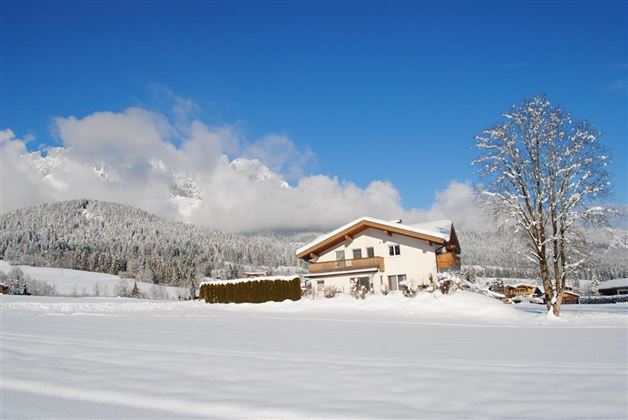 A snow-covered landscape with a modern house in the foreground. In the background, the snow-capped mountains rise under a blue sky.
