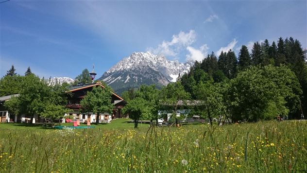 Een groene weide met kleurrijke bloemen en een traditioneel gebouw. Op de achtergrond rijzen besneeuwde bergen op onder een heldere blauwe lucht.