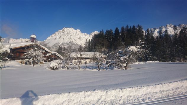 A winter landscape with snow-covered fields and mountains in the background. In the foreground stands a traditional wooden house surrounded by snow-covered trees.
