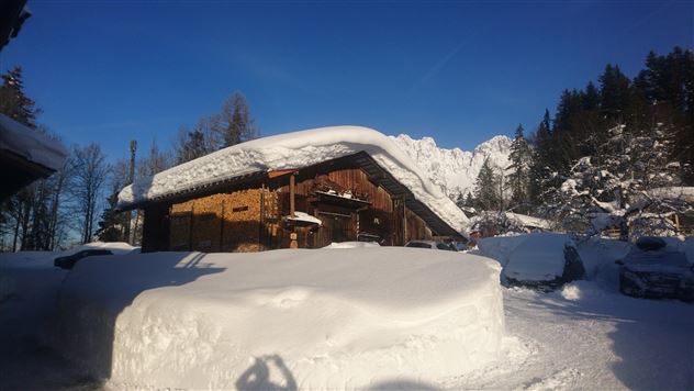 A snowy landscape with a traditional wooden house and mountains in the background. The sky is clear and blue, creating a calm winter atmosphere.