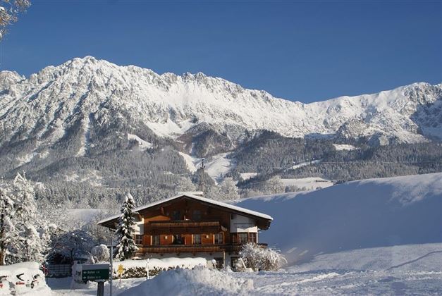 A charming wooden house in a snowy winter landscape. In the background, majestic mountains rise under a clear blue sky.