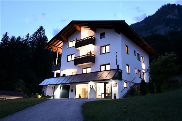 Ein modernes Haus im Alpenstil, das abends beleuchtet ist. Umgeben von Bäumen und Bergen, strahlt es eine einladende Atmosphäre aus.