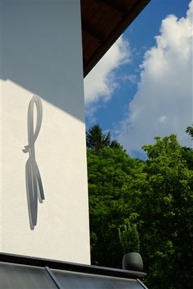 Ein modernes Gebäudewand mit einem stilisierten Logo. Im Hintergrund sind grüne Bäume und ein blauer Himmel mit einigen Wolken zu sehen.