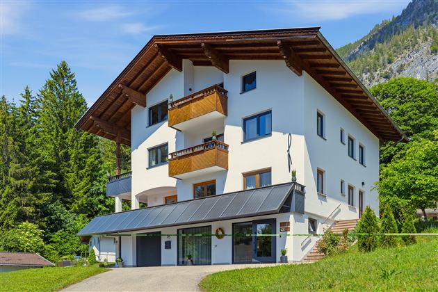 Ein modernes Haus mit Holzverzierungen und mehreren Balkonen. Es ist von Bäumen und grüner Landschaft umgeben.
