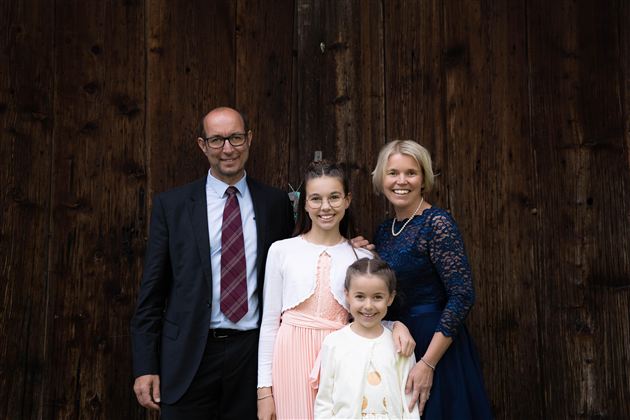 Eine Familie steht vor einer Holzwand. Sie lächeln freundlich und tragen festliche Kleidung.