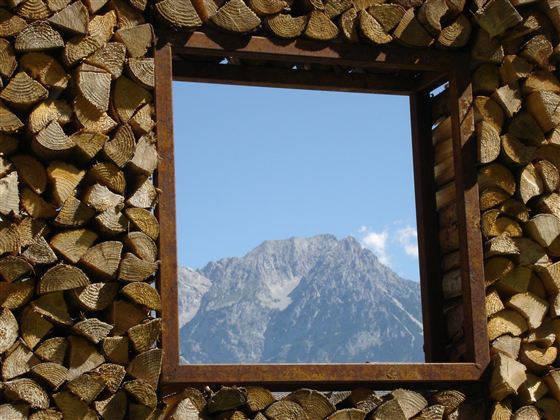 Ein Fensterrahmen aus Holzstämmen zeigt einen Blick auf einen majestätischen Berg und den blauen Himmel. Die Szene vermittelt Ruhe und Naturnähe.