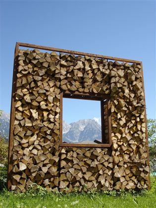 Een houten frame met een rechthoekige uitsnede, gevuld met houtblokken. Op de achtergrond zijn bergen en een heldere blauwe lucht te zien.