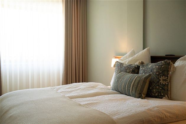 Een gezellige slaapkamer met een uitnodigend bed en zachte kussens. Het zachte licht van de ramen creëert een ontspannen sfeer.