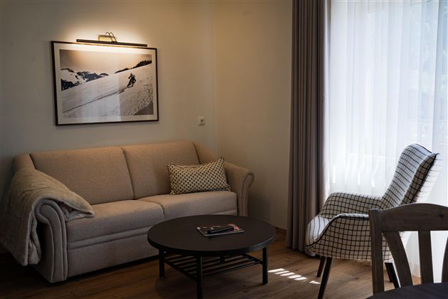 Een gezellige woonkamer met een lichte bank en een moderne fauteuil. Een ronde tafel en een groot raam zorgen voor veel daglicht.